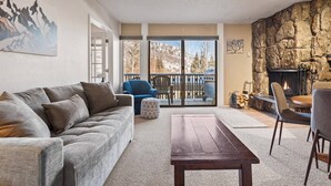 A living room with a sofa, a coffee table, a fireplace, and a view of mountains through large windows. - Snowmass Base Village Retreat. Great View W/ Balcony! Grill/Hot Tub/Pool/Parking. Monthly Rates! (Snowmass Village)