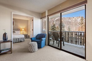 Une chambre d’hôtel avec un lit, un fauteuil bleu, un pouf à motifs et une grande porte-fenêtre coulissante donnant sur un balcon avec vue sur des arbres et des montagnes enneigés.