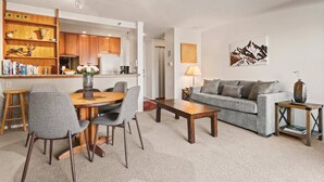 A modern living room with a grey sofa, wooden coffee table, and a dining area with a wooden table and grey chairs. - Snowmass Base Village Retreat. Great View W/ Balcony! Grill/Hot Tub/Pool/Parking. Monthly Rates! (Snowmass Village)