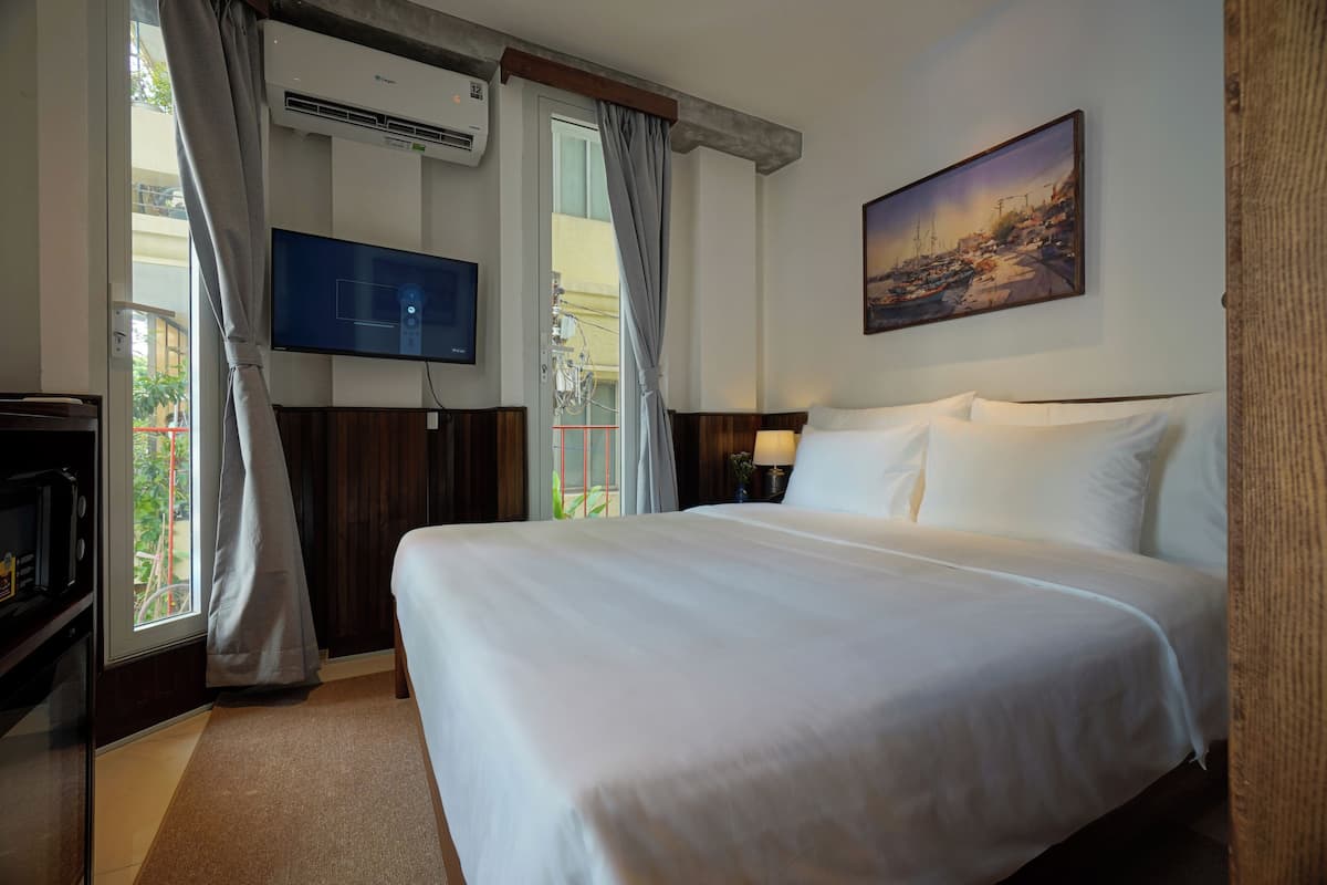 A hotel room with a large bed, a flat-screen TV mounted on the wall, a painting above the bed, and a window with curtains.
