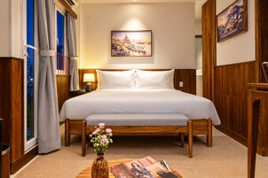 Habitación de hotel con una cama de madera, una mesita de noche con una lámpara, un jarrón de flores y una ventana con cortinas.