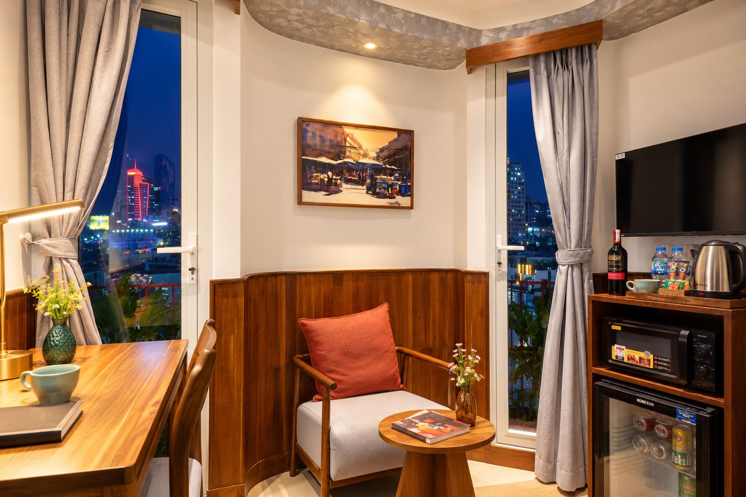 Une chambre d’hôtel avec vue sur la ville, un chevet en bois, un canapé, une table avec un livre, une petite télévision et un mini-bar.