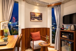 A hotel room with a city view, a wooden headboard, a sofa, a table with a book, a small TV, and a minibar. - Nexus House Saigon Central - District 1 (Ho Chi Minh City)