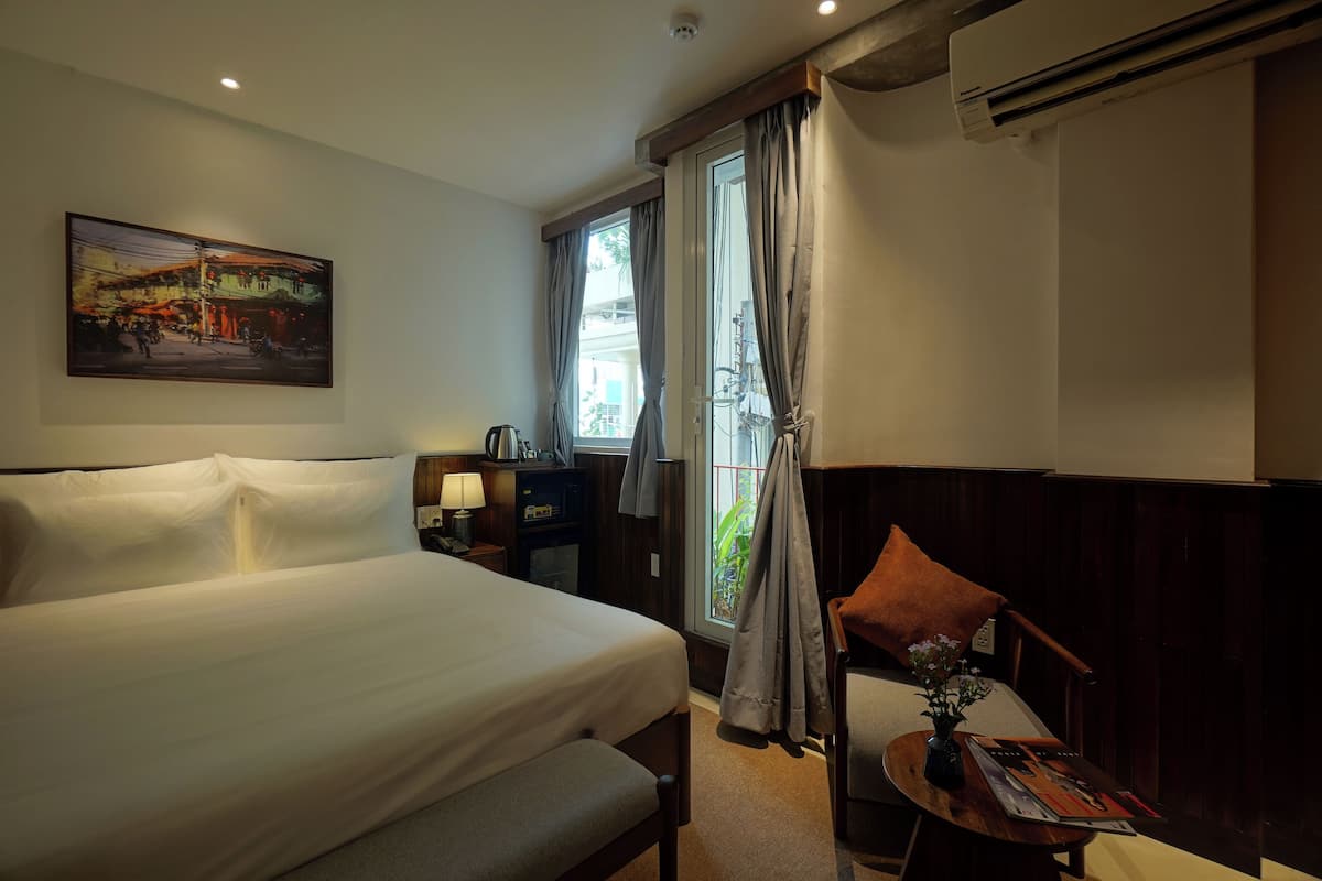 A hotel room with a large bed, a desk with a chair, a window with curtains, a painting on the wall, and an air conditioning unit.