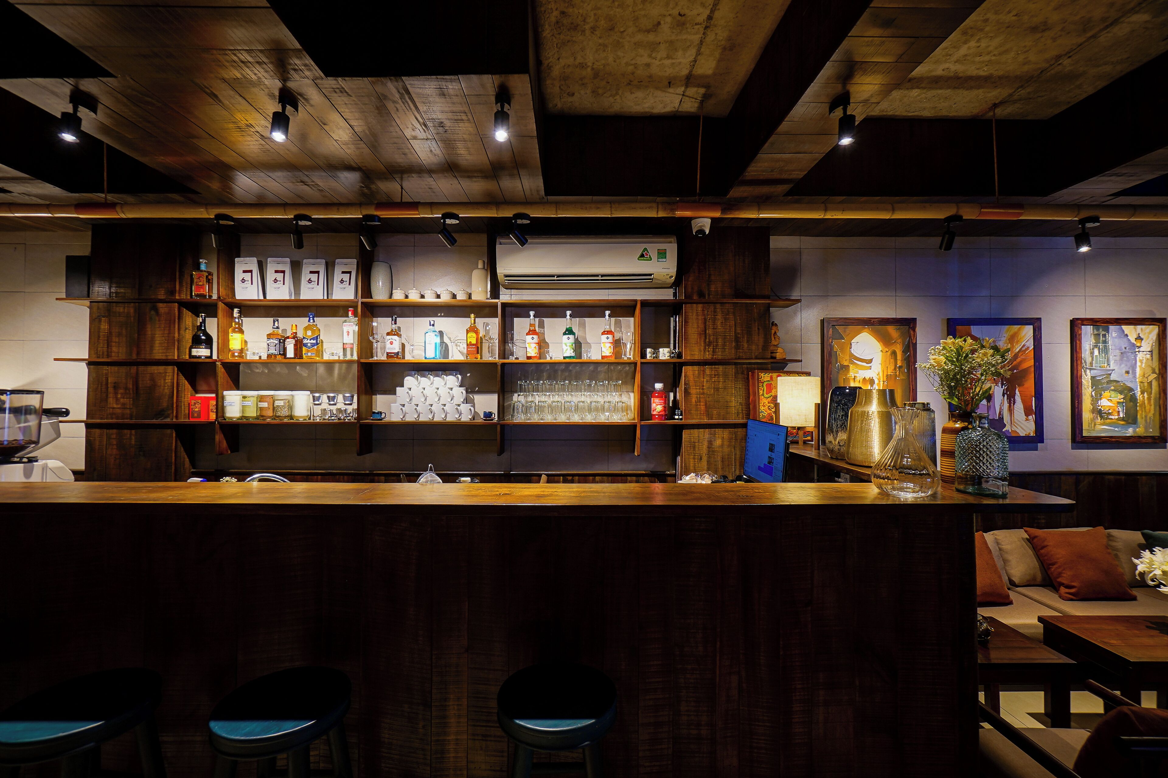 Una zona de bar con estantes de madera llenos de botellas y vasos, una barra y taburetes.