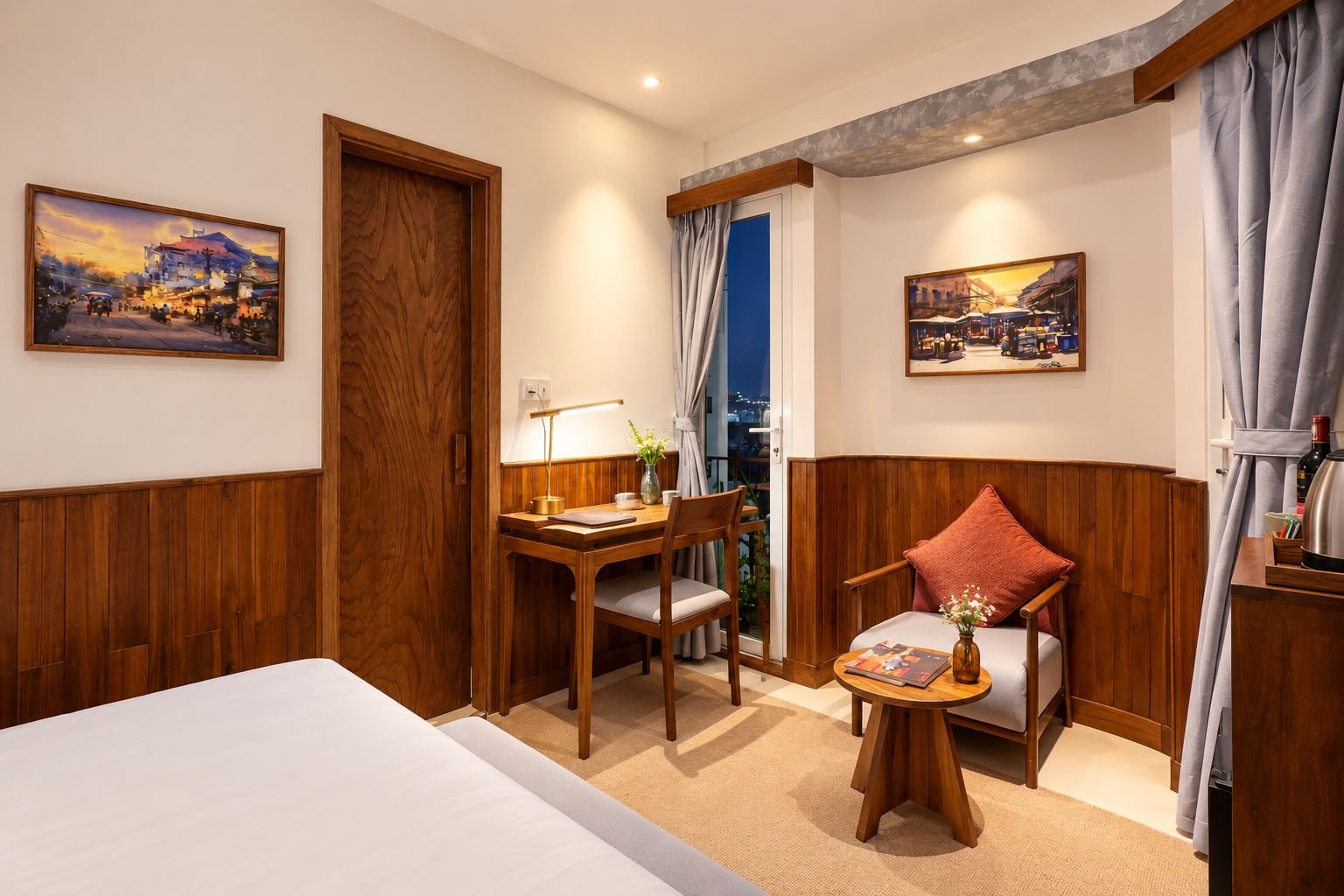 A hotel room with a bed, a desk with a lamp, a chair, a small table with a vase, and two paintings on the walls.