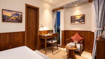 A hotel room with a bed, a desk with a lamp, a chair, a small table with a vase, and two paintings on the walls.