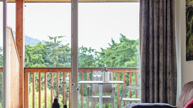 En balkon med gelænder af træ, en fletstol, et lille bord med to vaser, samt udsigt til grønne områder og bjerge.
