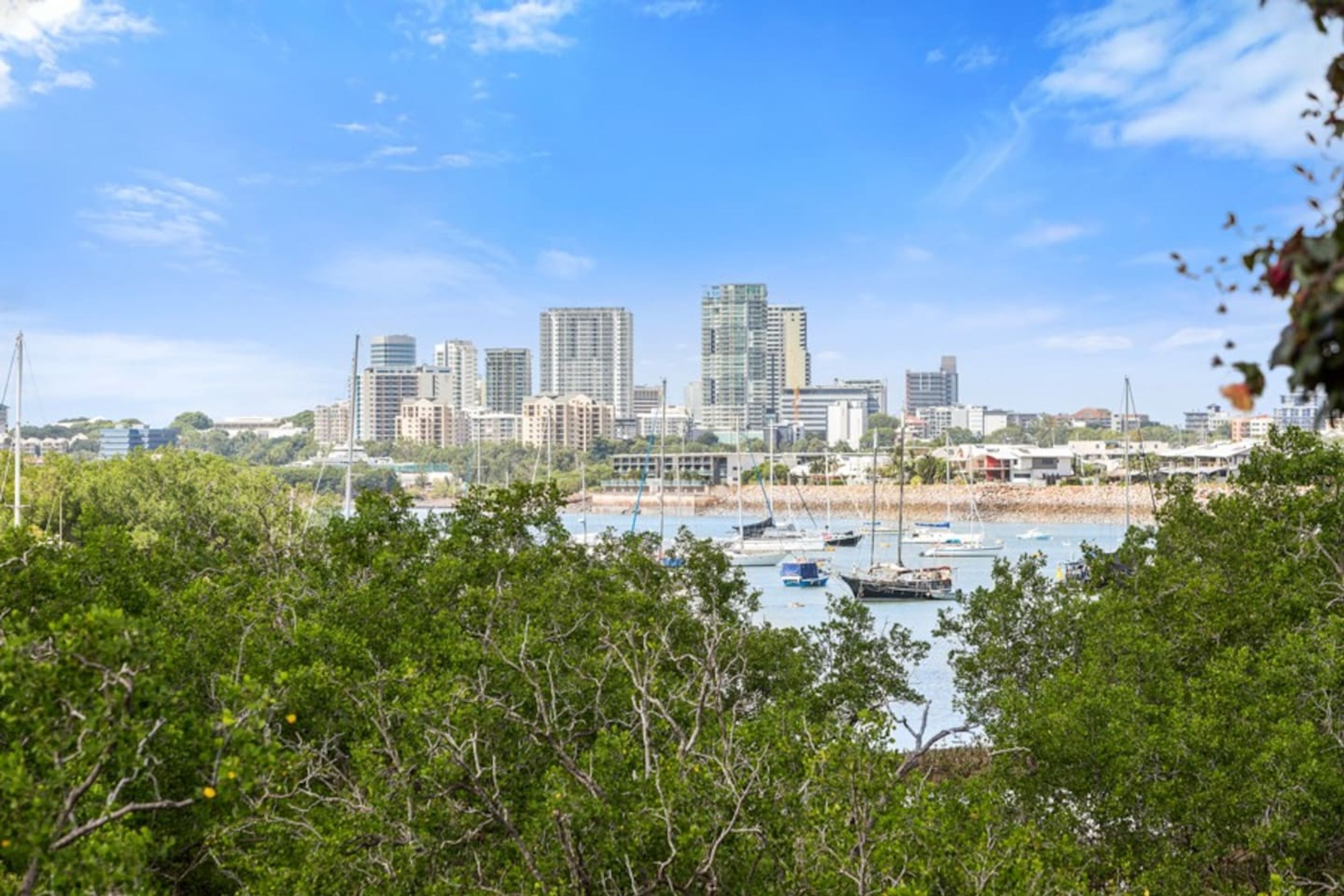A coastal cityscape with modern high-rise buildings, a marina with boats, and lush greenery in the foreground.