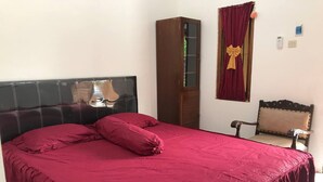 Ein Bett mit dunklem Kopfteil, ein hoher Holzkasten, ein Stuhl und ein Fenster mit rotem Vorhang.