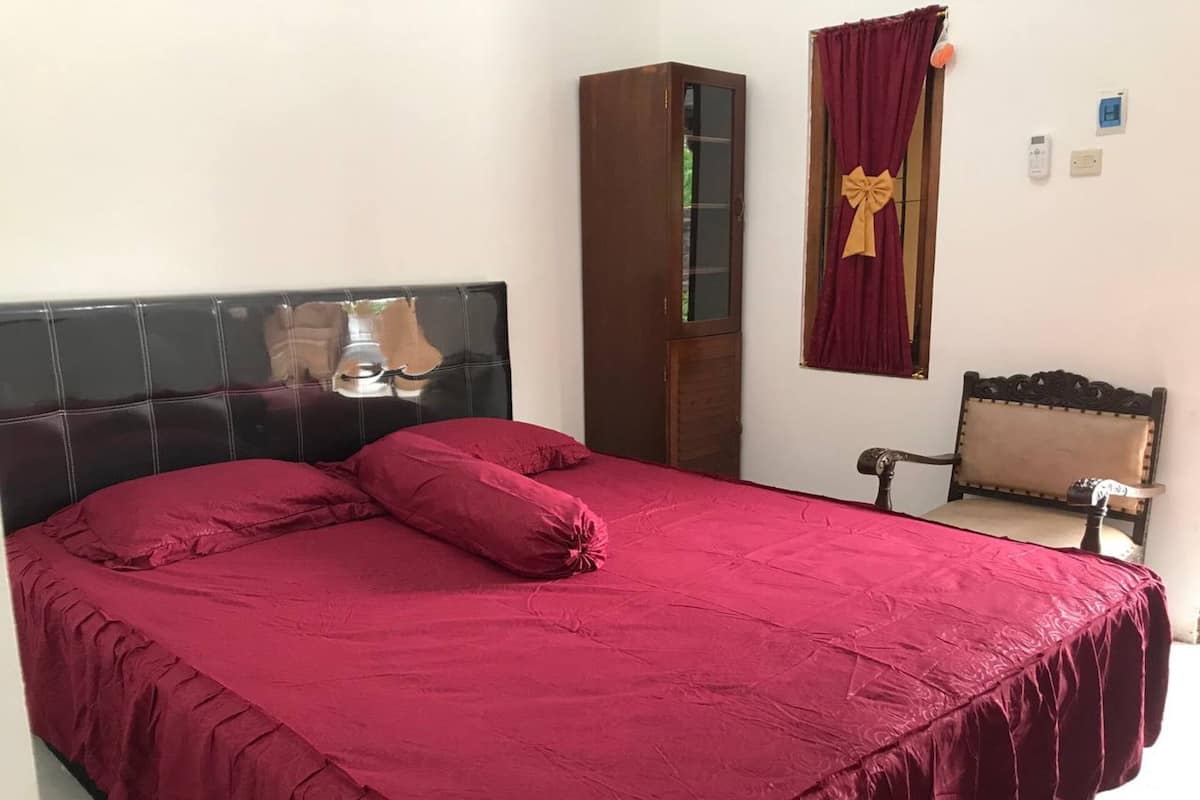 Ein Bett mit dunklem Kopfteil, ein hoher Holzkasten, ein Stuhl und ein Fenster mit rotem Vorhang.