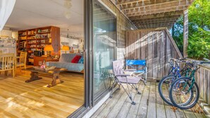 Una sala de estar acogedora con sofá, mesa de centro y estantería, un comedor con sillas y una terraza de madera con bicicletas.