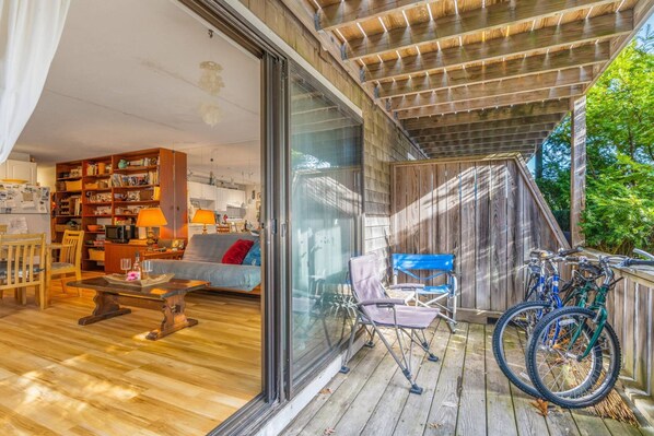 A cozy living room with a sofa, coffee table, and bookshelf, a dining area with chairs, and a wooden deck with bicycles.