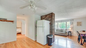 Amplia sala de estar con piso de madera, refrigerador blanco, pared de ladrillo decorativa, ventilador de techo y zona de comedor con mesa y sillas.