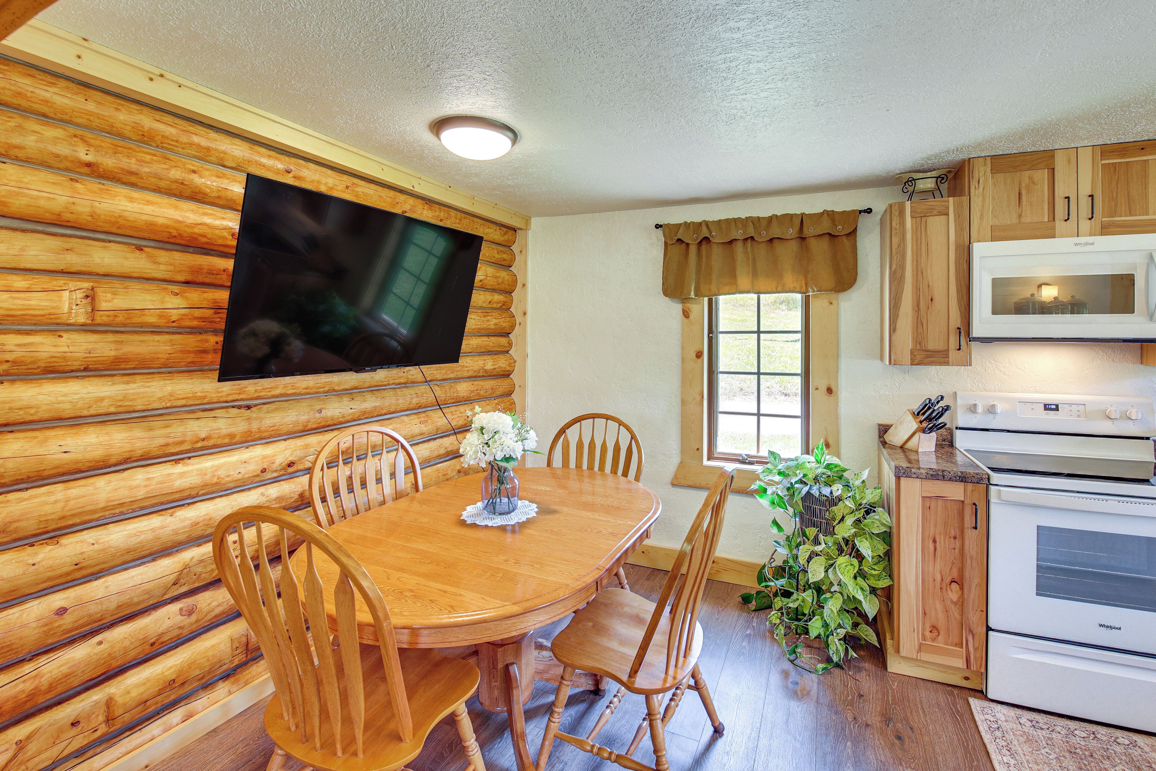 A wooden interior with a dining table, chairs, a television mounted on the wall, a window with curtains, and a microwave in the kitchen.