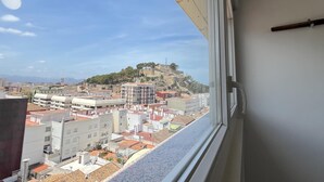 Vista desde una ventana que da a un paisaje urbano con edificios, una colina con una estructura similar a un castillo y un cielo despejado.