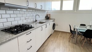 Cocina moderna con gabinetes blancos, encimera de granito, estufa a gas, lavabo y lavadora. Cuenta con una zona de comedor con mesa y sillas de vidrio.