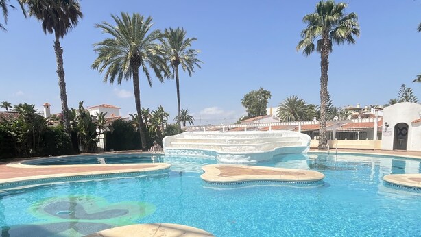 A swimming pool with a central island, surrounded by palm trees and buildings. - Flat El Retiro 4 people with swimming pool. (Dénia)