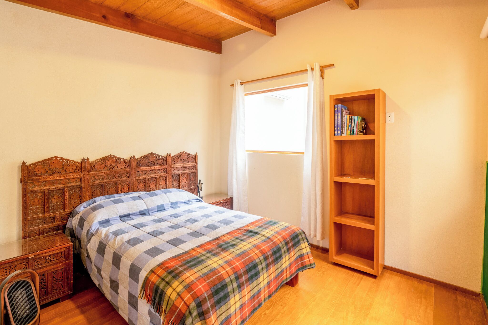 A bedroom with a wooden headboard, a checkered bedspread, a wooden bookshelf, and a window with curtains.