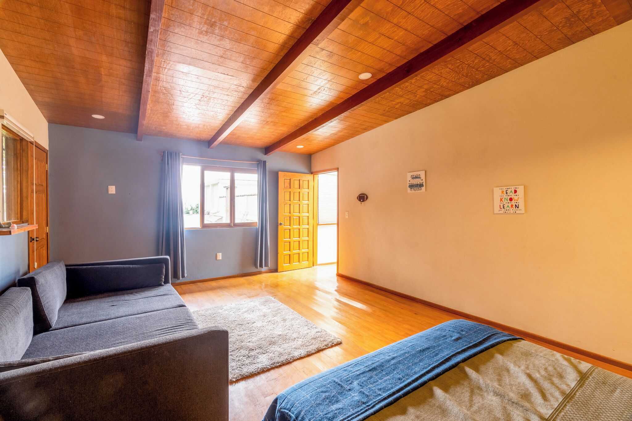 Ein geräumiger Raum mit Holzbalkendecke, einem grauen Sofa, einer blauen Decke und einer Holztür.