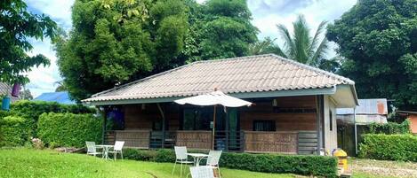 A small building with a tiled roof, surrounded by lush greenery and trees. There are outdoor tables and chairs set up for dining.