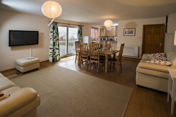A living room with a TV, a dining area with wooden chairs, a sofa, and a kitchen in the background.