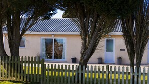 A single-story building with a tiled roof, a white door, and a small window. There is a wooden fence in the foreground, and trees are visible behind it. - Drws Nesa - sleeps 6 guests  in 3 bedrooms (Pwllheli)