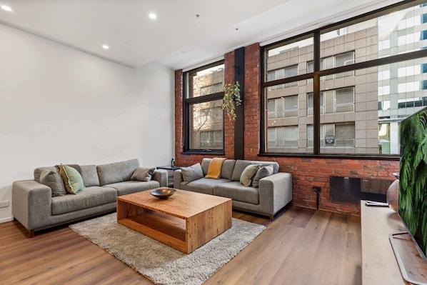 A modern living room with two grey sofas, a wooden coffee table, and large windows with a brick wall.