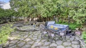 A stone patio with a glass table and chairs, surrounded by lush greenery and a stone wall. - 5BR Chalet with Hot Tub & Lake Access (Mont-Blanc)