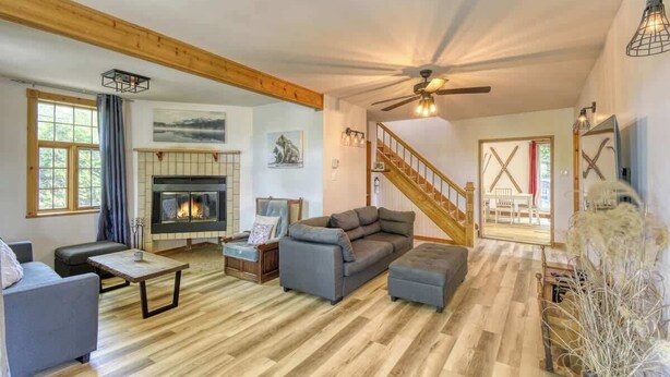 A spacious living room with a fireplace, wooden beams, a staircase, and a view of the outdoors. - 5BR Chalet with Hot Tub & Lake Access (Mont-Blanc)
