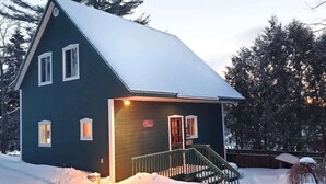 Une maison d’un vert foncé, avec un toit recouvert de neige, un porche à l’avant et un escalier menant à l’entrée.