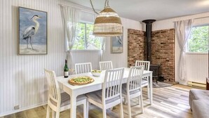 Un coin repas avec une table et des chaises blanches, un luminaire suspendu en osier, une cheminée en briques et un tableau représentant un oiseau, encadré, accroché au mur.