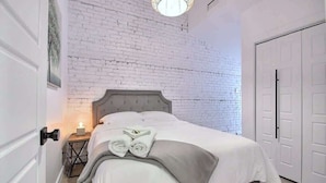 Un dormitorio con pared de ladrillo blanco, cama con cabecera gris, mesita de noche con lámpara y puerta blanca.