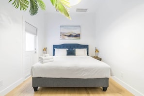 Une chambre à coucher avec un lit, des murs blancs, une tête de lit bleue, un tableau et une plante.
