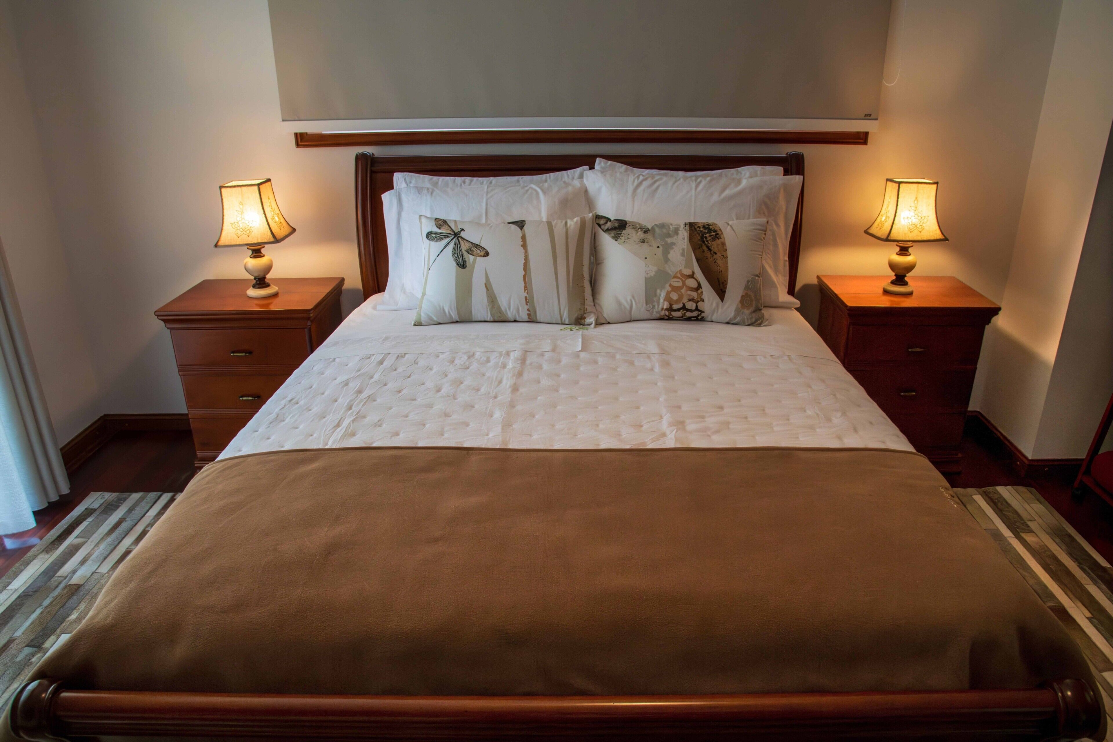 Cama bem arrumada com colcha marrom, duas mesas de cabeceira com luminárias e almofadas decorativas.