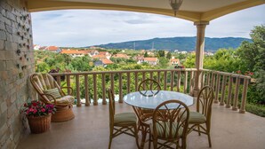 Een balkon met rieten stoelen, een glazen tafel en uitzicht op een dorp en bergen.
