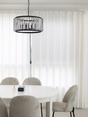 Un coin repas avec une table ronde, quatre chaises, un lustre moderne et de fines rideaux blancs.