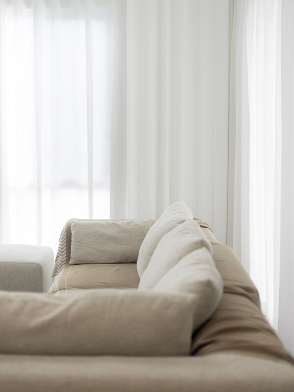 A room with a beige sofa and a white pillow, a window with sheer curtains, and a white wall.
