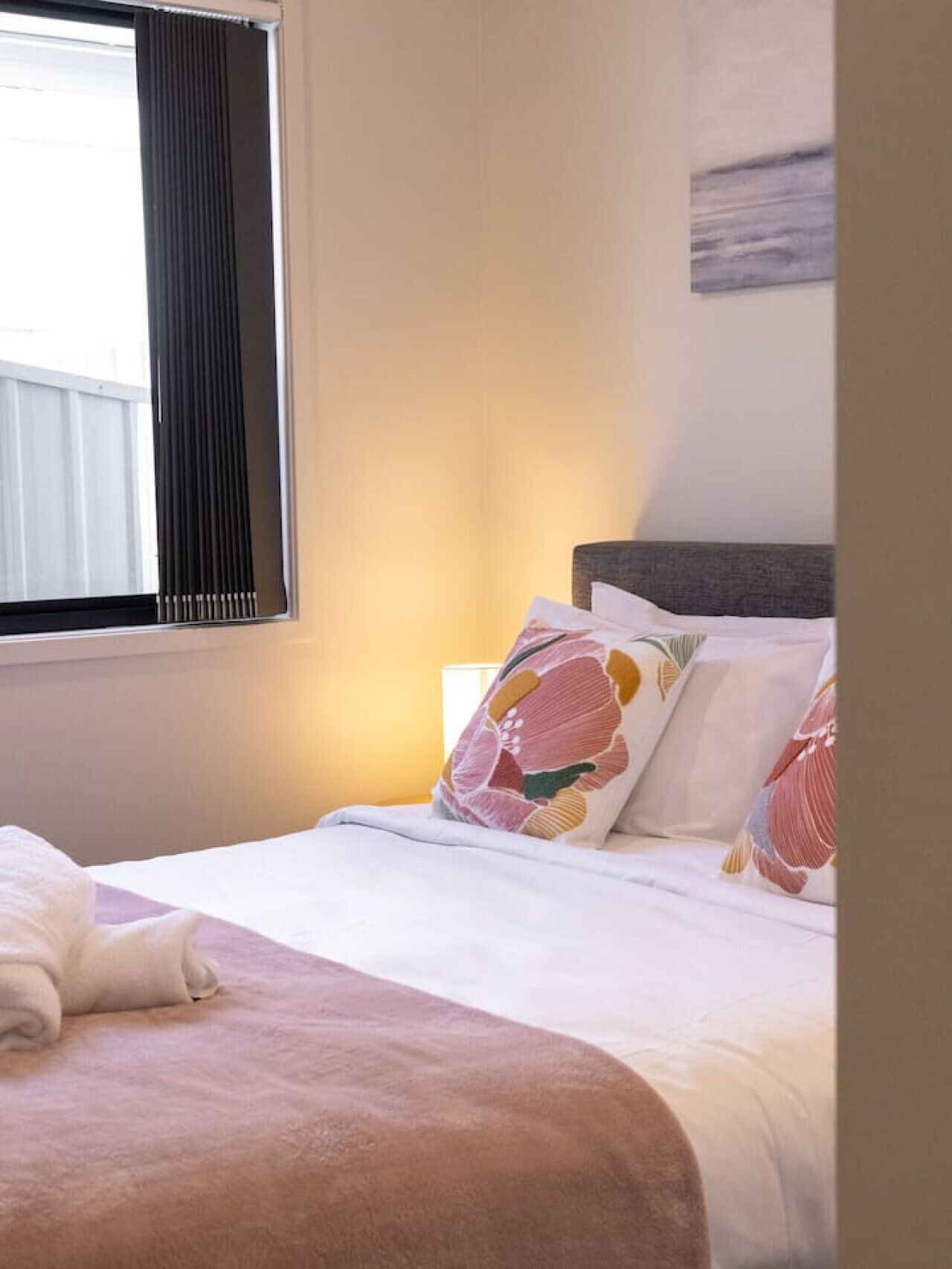 Ein ordentlich bezogenes Bett mit einer rosa Decke und blumigen Kissen, ein Fenster mit schwarzen Vorhängen und eine Wand mit einem schlichten Gemälde.
