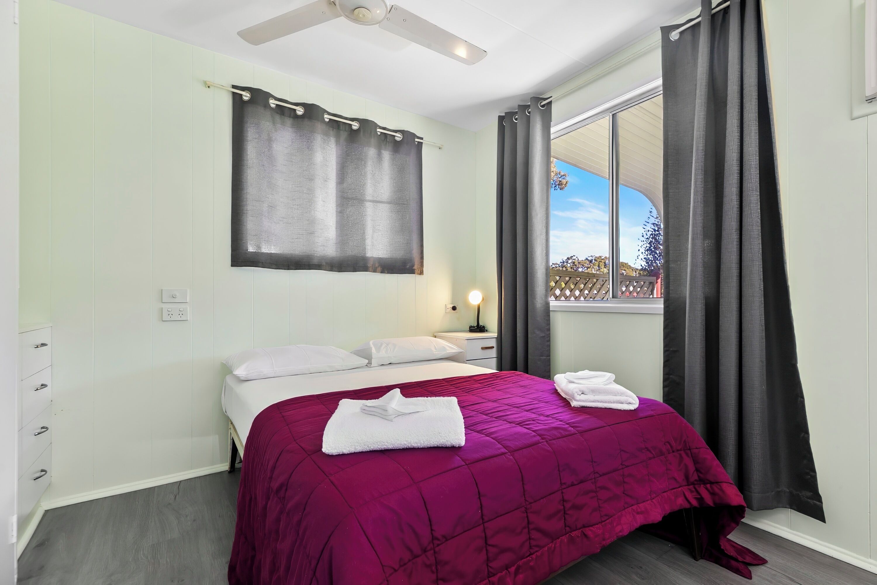 Une chambre à coucher avec un lit, une table de chevet, une fenêtre avec des rideaux et un ventilateur de plafond.