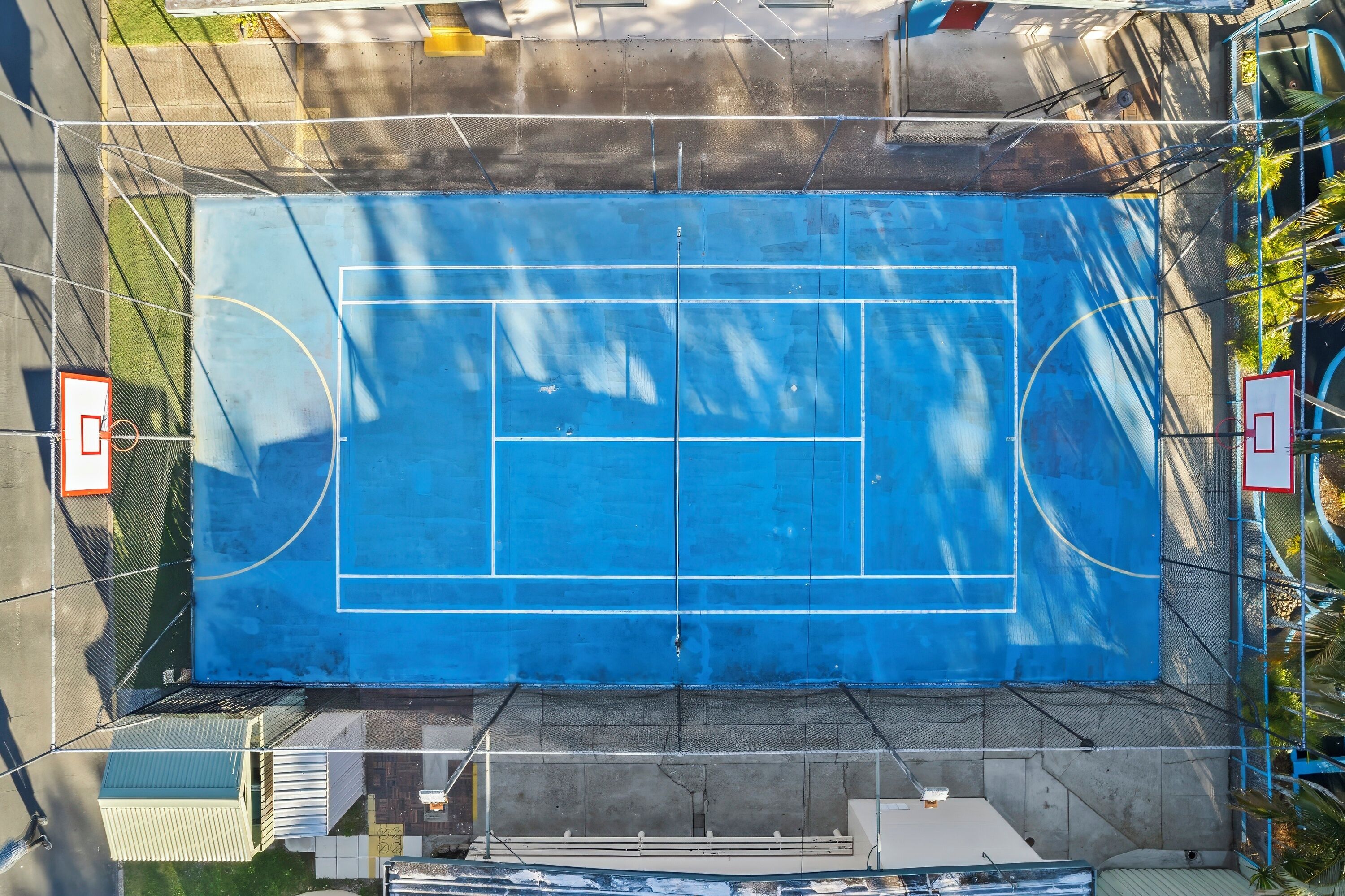 Une vue aérienne d’un terrain de basket-ball bleu, avec un panier et un filet, entouré d’une clôture et d’arbres.