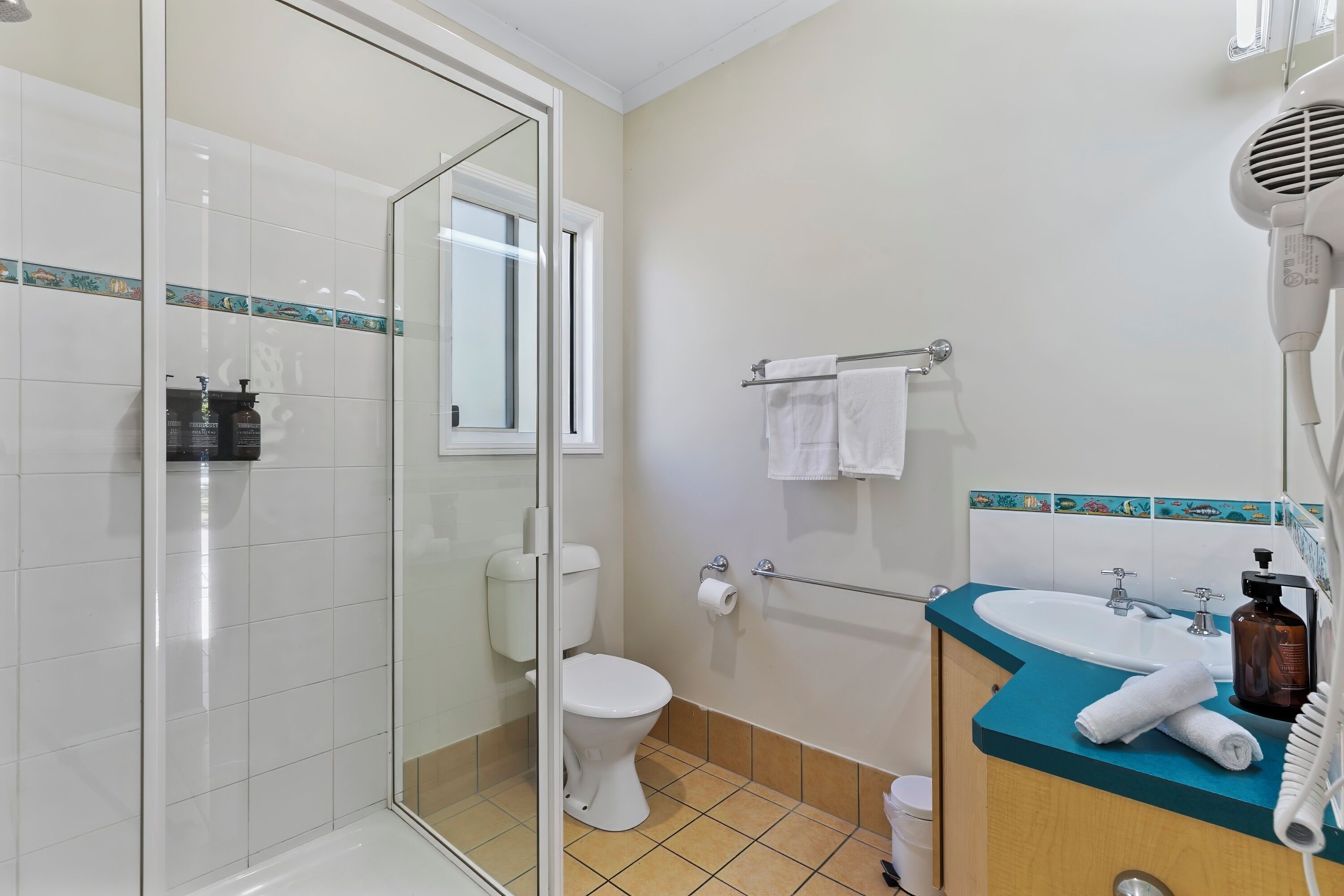 A bathroom with a glass shower enclosure, a toilet, a sink with a towel rack, and a hairdryer on the counter.