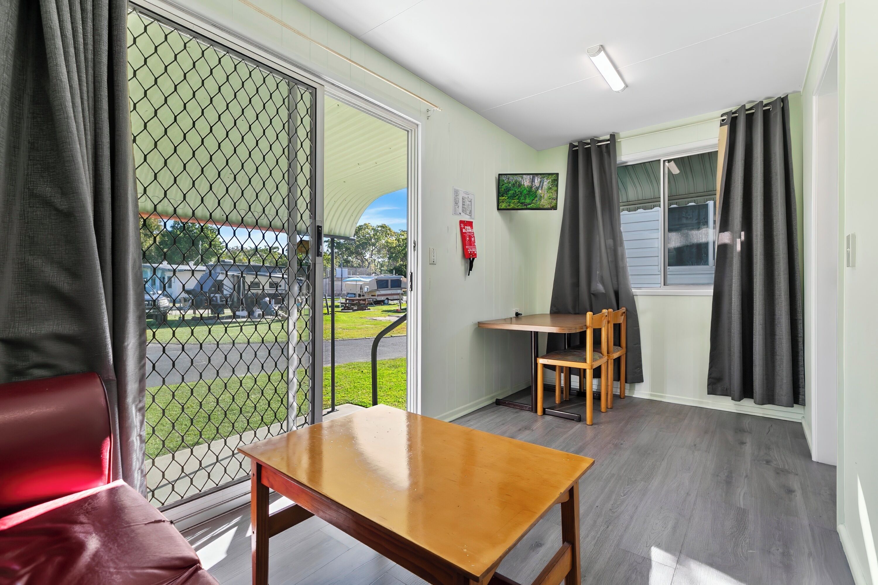 A room with a wooden table and chairs, a red couch, a large window with a view of a parking lot, and a fire extinguisher on the wall.
