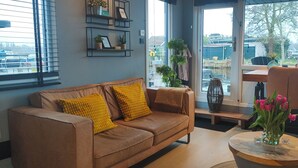 Un salon moderne avec un canapé en cuir marron, des coussins jaunes, une table basse en bois ornée d’un vase rempli de fleurs roses, et une vue sur un parking à travers de grandes fenêtres.