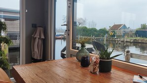 Une table en bois, ornée d’un vase, d’une plante en pot et d’un sac de noix, est placée près d’une fenêtre donnant sur un canal où l’on peut voir des bateaux et des bâtiments.