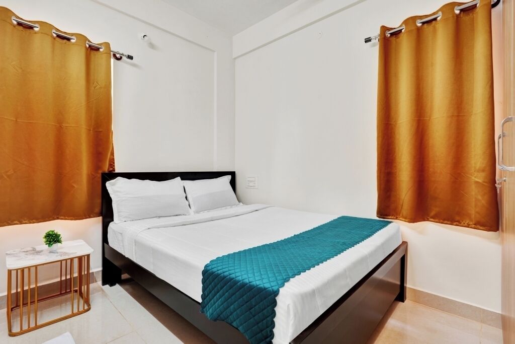 A neatly made bed with white linens and a teal blanket, a small bedside table with a plant, and a window with a curtain.