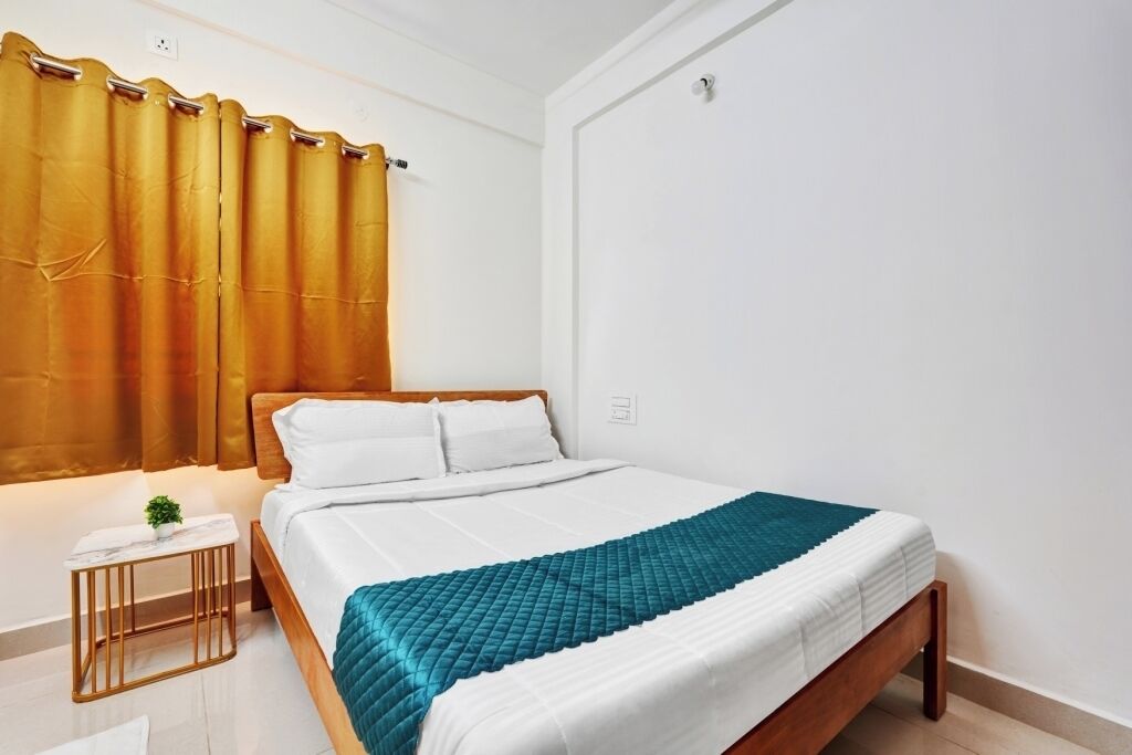 A neatly made bed with a white and teal quilt, a small bedside table with a plant, and a golden curtain.