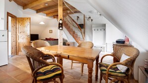 Un coin repas avec une table en bois et des chaises en osier, un escalier muni d’un filet de sécurité, et une cuisine en arrière-plan.