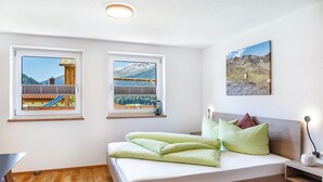 A bedroom with a bed, two windows with mountain views, a bedside table, and a lamp. - Apartment 'Berghoamat Nauders' with Mountain View, Private Terrace and Wi-Fi (Nauders)