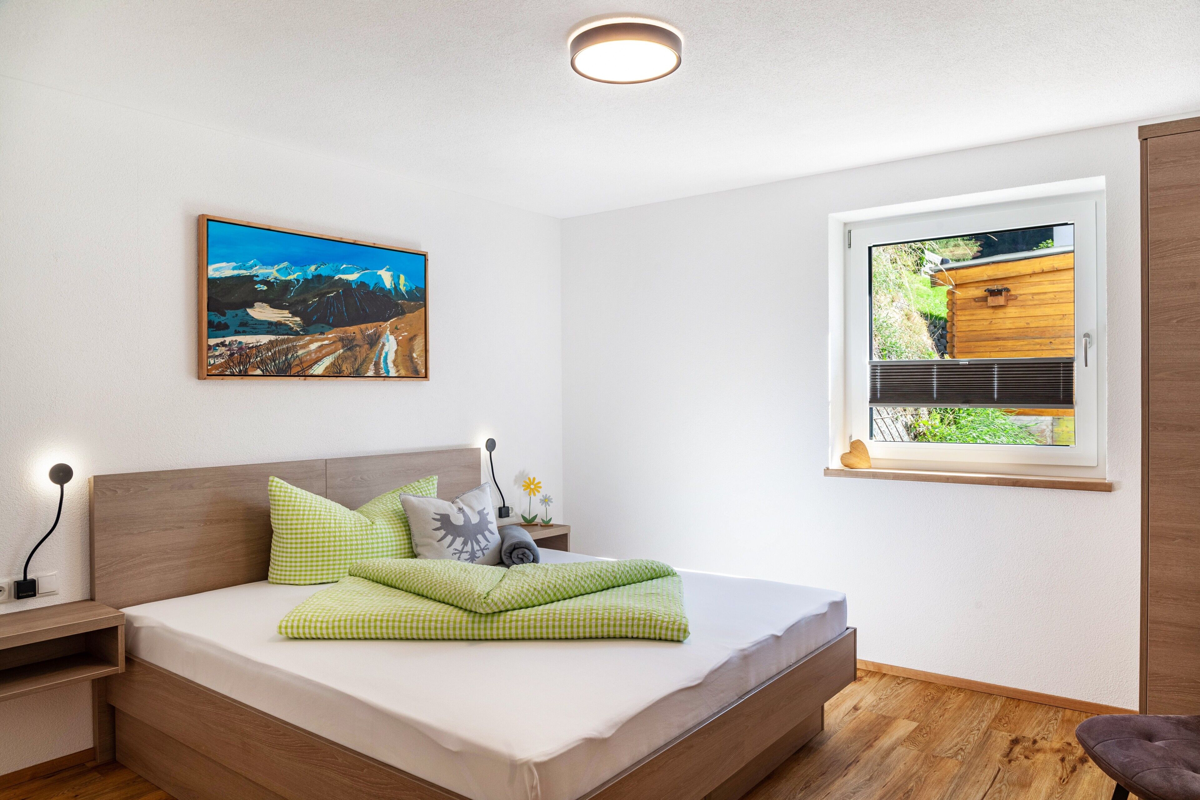 Un dormitorio con una cama, una mesita de noche, un cuadro colgado en la pared, una ventana con vistas y una lámpara en el techo.